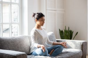 Meditation for Addiction Recovery, pretty young woman sitting cross-legged on couch at home, meditating - types of meditation