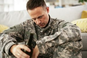 man in camoflauge at home sitting on floor drinking a bottle of beer - veterans and addiction