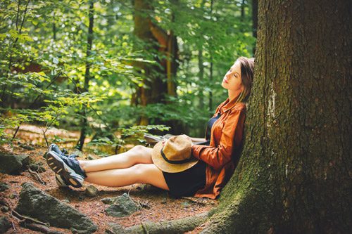 young woman in hiking clothes sitting, leaned against a large tree in a coniferous forest - stress in recovery
