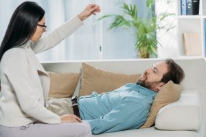 man lying on psychotherapist's couch while she uses a pendant to hypnotize him - Trauma-Informed Hypnotherapy