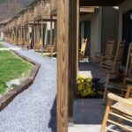 Row of wooden rocking chairs on a covered porch outside a rustic building, with a gravel path and green lawn in a peaceful outdoor setting.