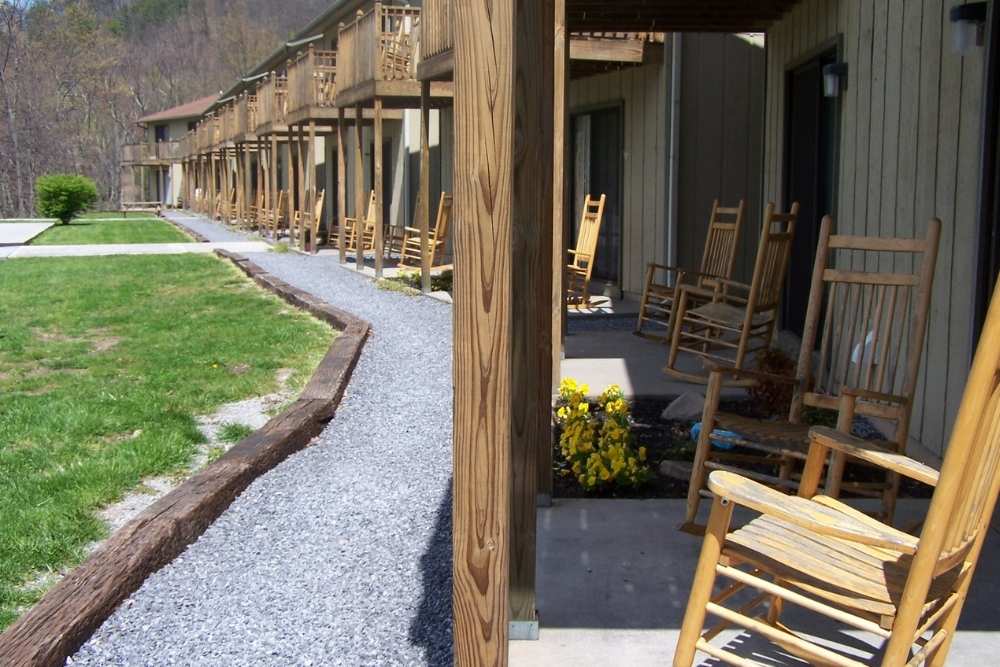 Row of wooden rocking chairs on a covered porch outside a rustic building, with a gravel path and green lawn in a peaceful outdoor setting.