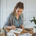 Woman sitting at a table painting leaves on paper with watercolors, surrounded by art supplies