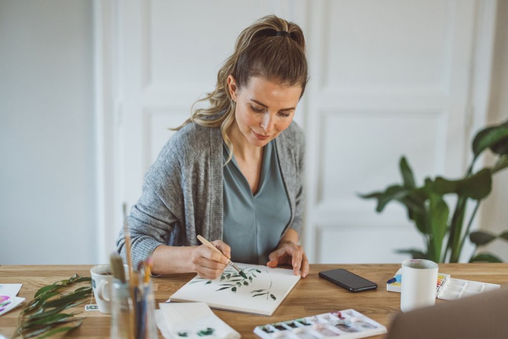 Woman sitting at a table painting leaves on paper with watercolors, surrounded by art supplies