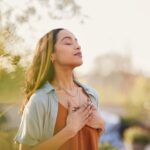 Woman standing outdoors with eyes closed and hands on her chest, appearing calm and centered, symbolizing mindfulness and inner peace.