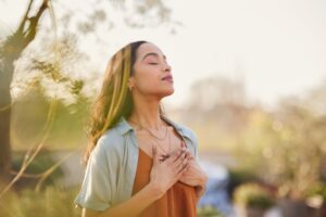 Woman standing outdoors with eyes closed and hands on her chest, appearing calm and centered, symbolizing mindfulness and inner peace.