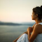Woman sitting outdoors at sunset, gazing calmly at a scenic view of water and distant mountains, embracing a peaceful moment in nature.
