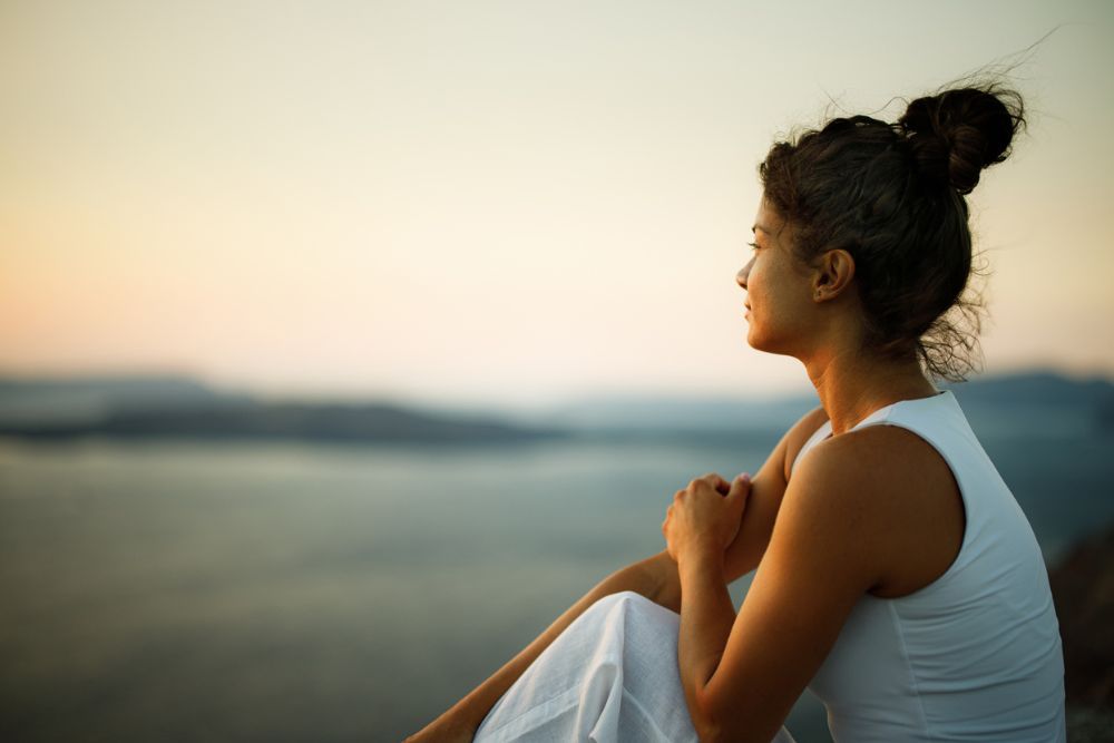 Woman sitting outdoors at sunset, gazing calmly at a scenic view of water and distant mountains, embracing a peaceful moment in nature.
