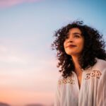 Young woman with curly hair smiles peacefully while gazing at the colorful sunset sky, reflecting a calm and hopeful mood.