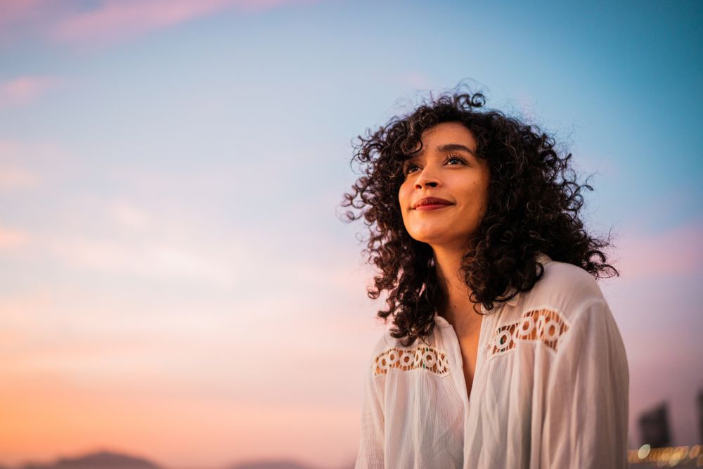 Young woman with curly hair smiles peacefully while gazing at the colorful sunset sky, reflecting a calm and hopeful mood.