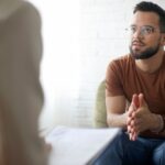 A young man with glasses and a beard sits on a couch, leaning forward and talking attentively to a therapist holding a clipboard. The setting is bright with a white brick wall in the background.