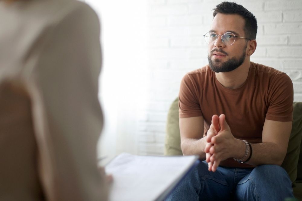 A young man with glasses and a beard sits on a couch, leaning forward and talking attentively to a therapist holding a clipboard. The setting is bright with a white brick wall in the background.