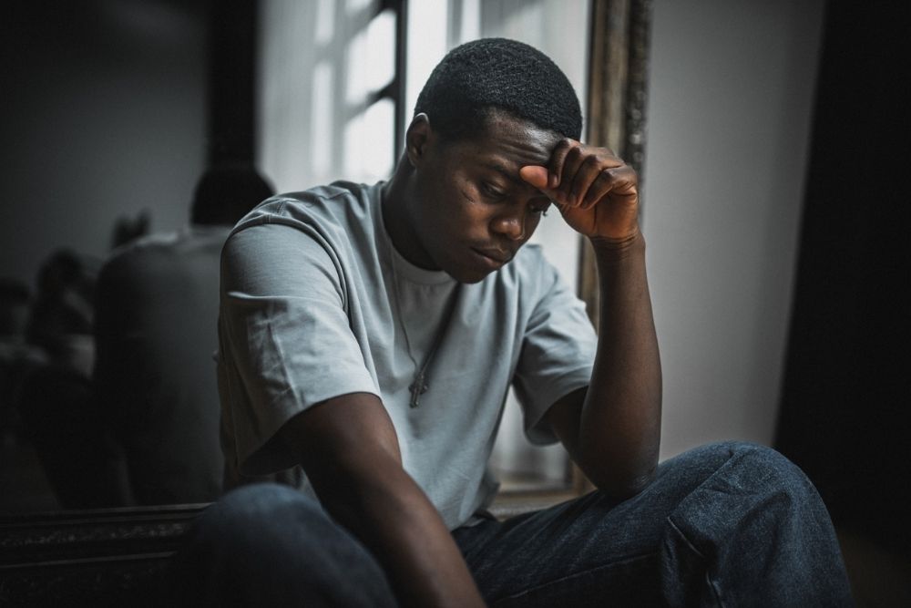 Man sitting indoors with his head resting on his hand, looking down with a serious or reflective expression, suggesting emotional distress or deep thought.