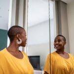 A smiling woman in a yellow top looks at her reflection in the mirror, appearing confident and happy in a bright, modern room.