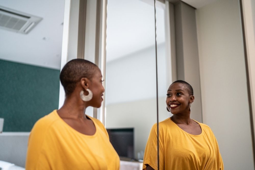 A smiling woman in a yellow top looks at her reflection in the mirror, appearing confident and happy in a bright, modern room.