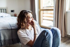 A woman sits on the floor beside a bed, resting her hand near her mouth and looking thoughtful or concerned toward the window.