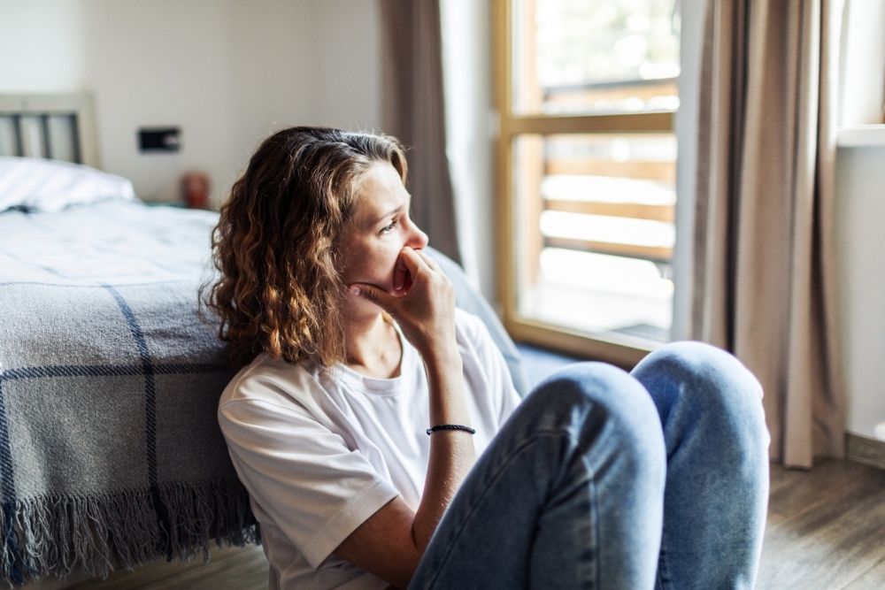 A woman sits on the floor beside a bed, resting her hand near her mouth and looking thoughtful or concerned toward the window.