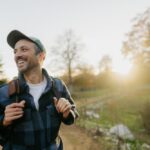 Man smiling while hiking outdoors on a sunny day, wearing a cap and backpack, surrounded by trees and soft golden light.