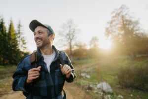 Man smiling while hiking outdoors on a sunny day, wearing a cap and backpack, surrounded by trees and soft golden light.