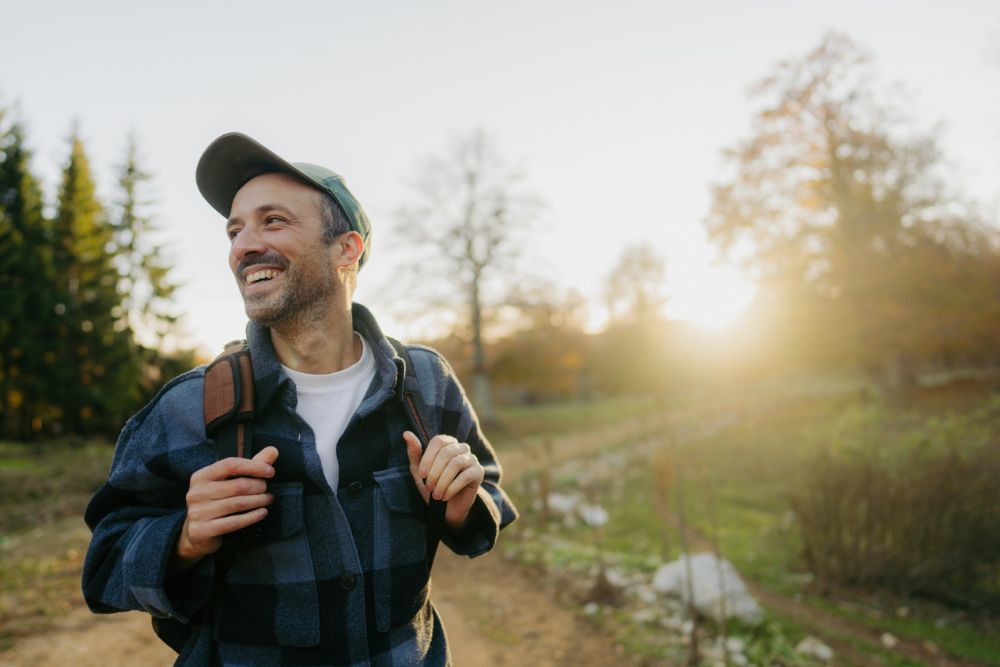 Man smiling while hiking outdoors on a sunny day, wearing a cap and backpack, surrounded by trees and soft golden light.