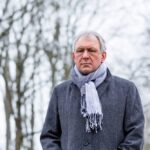 An older man standing outdoors on a cold day, wearing a gray coat and a scarf, looking serious with bare winter trees in the background.