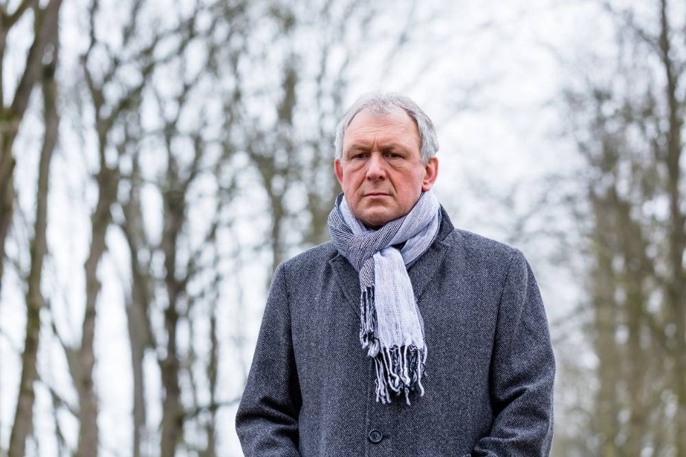 An older man standing outdoors on a cold day, wearing a gray coat and a scarf, looking serious with bare winter trees in the background.