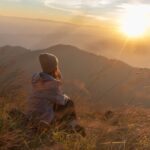 Person wearing a beanie sitting in tall grass on a hillside, peacefully watching the sunrise over distant mountains, symbolizing reflection and purpose.