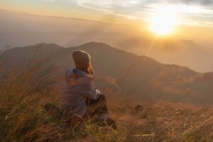 Person wearing a beanie sitting in tall grass on a hillside, peacefully watching the sunrise over distant mountains, symbolizing reflection and purpose.