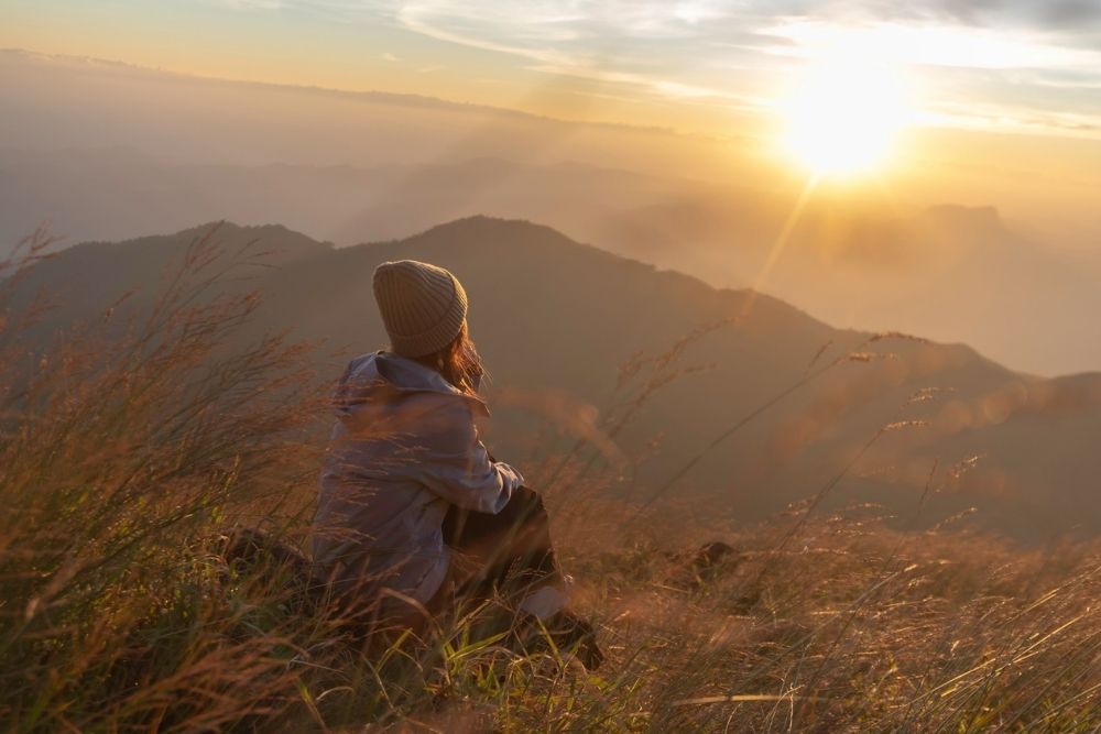 Person wearing a beanie sitting in tall grass on a hillside, peacefully watching the sunrise over distant mountains, symbolizing reflection and purpose.