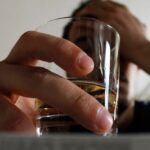 A person holds a glass of alcohol while resting their head in their hand, suggesting distress or overdrinking.