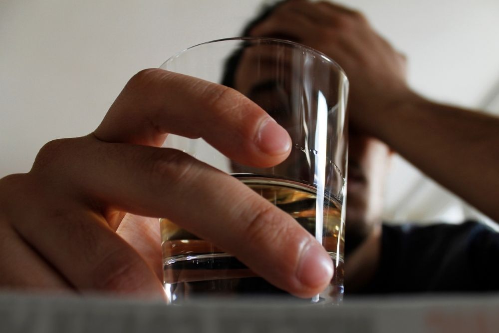 A person holds a glass of alcohol while resting their head in their hand, suggesting distress or overdrinking.