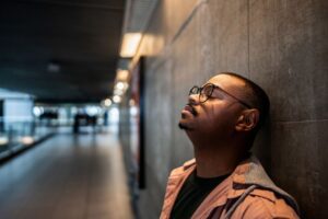 A man wearing glasses leans against a wall with his eyes closed, appearing tense or overwhelmed, symbolizing anxiety or emotional strain.