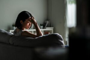 Woman sitting on a couch in a dimly lit room with her hand on her forehead, appearing deep in thought or emotionally distressed.