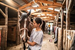 Woman gently holding and interacting with a horse inside a stable, suggesting a calm therapeutic or bonding moment.