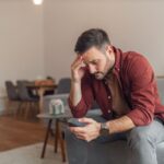 Relapse Treatment (1000 x 667 px) A man sitting on a couch holding his phone and resting his head in his hand, appearing worried or stressed.