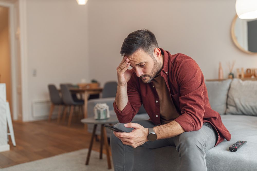 A man sitting on a couch holding his phone and resting his head in his hand, appearing worried or stressed.