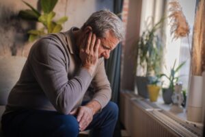 Older adult sitting by a window with head in hand, appearing overwhelmed or distressed in a quiet home environment.