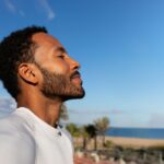 English Mountain – Strategies for Managing Stress in Addiction Recovery – Edited Strategies for Managing Stress in Addiction Recovery - A man in a white shirt stands outdoors with eyes closed, enjoying the sun. Palm trees and a beach in the background under a clear blue sky convey tranquility.