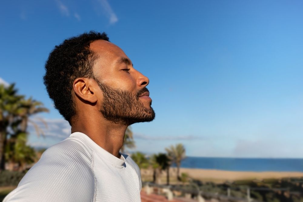 Strategies for Managing Stress in Addiction Recovery - A man in a white shirt stands outdoors with eyes closed, enjoying the sun. Palm trees and a beach in the background under a clear blue sky convey tranquility.