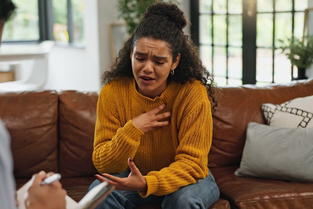 English Mountain - What Is Somatic Trauma Recovery and Can It Help You - Edited. A woman in therapy speaks emotionally to her therapist while sitting on a brown couch in a well lit room.